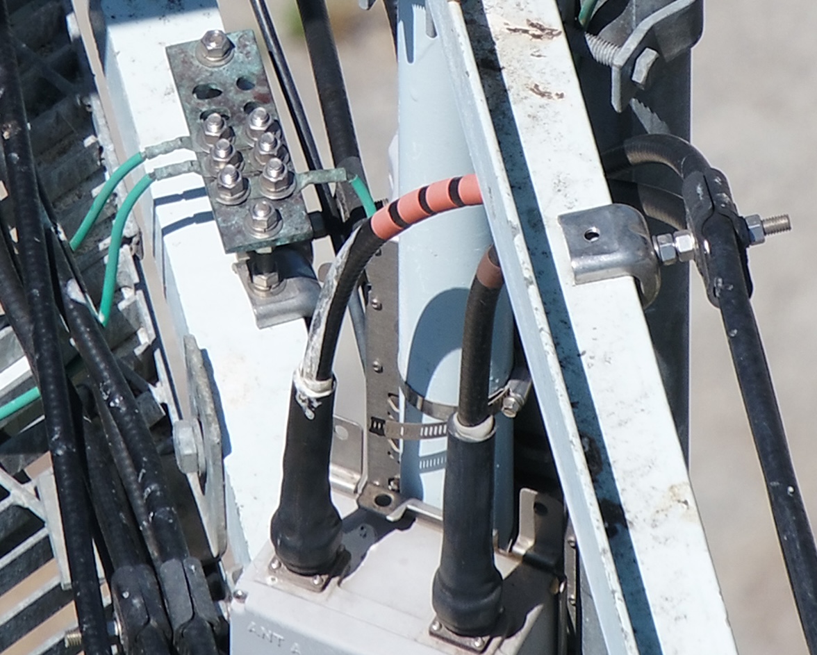 High-resolution inspection detail of cell-site hardware — a grounding bar with bolt terminals, green ground wires, color-banded coax jumpers connecting to antenna ports, and mounting clamps captured during an autonomous pass