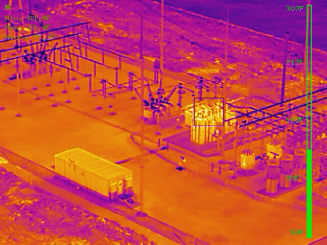 Aerial radiometric thermal capture of an electrical substation in the ironbow palette, with a temperature scale and measured hot-spot readings on transformer and bus components