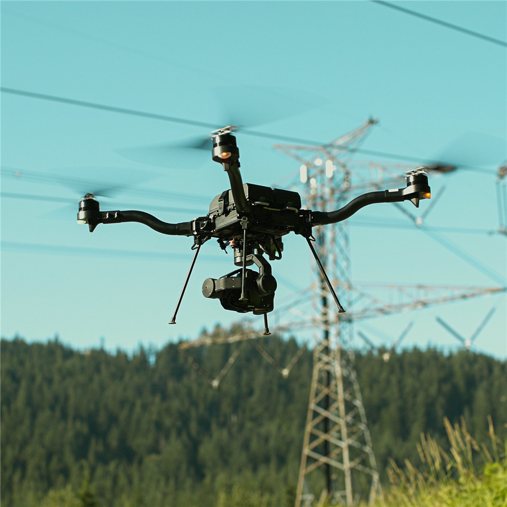 Freefly Astro drone with a gimbaled sensor payload flying beside a steel lattice transmission tower and conductors over forested terrain during autonomous corridor capture